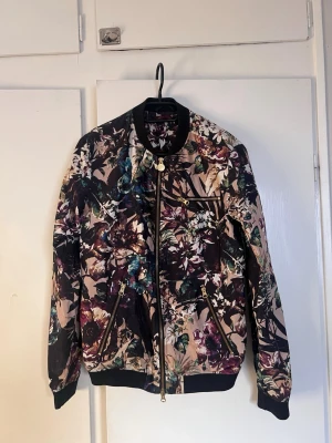 Blommig bomberjacka  - Snygg bomberjacka från nikolaj d'etoiles med färgstarkt blommönster i lila, grönt, beige och svart. Jackan har svarta muddar, guldfärgade dragkedjor och en cool bröstficka med zip. Perfekt för dig som vill sticka ut med din stil.