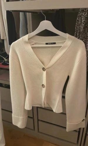 Vit kofta från Gina Tricot - Stilren vit kofta från Gina Tricot med v-ringning och stora knappar framtill. Koftan har långa ärmar med breda muddar och en något croppad passform. Perfekt att slänga över en topp för en enkel men snygg look.