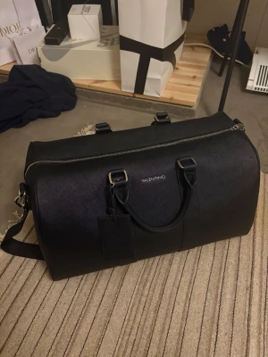 Svart weekendbag från Valentino - Snygg svart weekendbag från Valentino i ett tåligt konstläder med struktur. Väskan har dubbla handtag, avtagbar axelrem och silverfärgade metalldetaljer. Perfekt storlek för kortare resor och stilren design med diskret logga framtill.