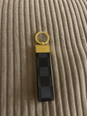 Vuitton nyckelring i canvas - Snygg nyckelring från  Vuitton i svart och grå canvas med rutmönster, detaljer i guldmetall och brunt läder. Klassisk -logga på metallfästet och präglad text på lädret. Perfekt accessoar för att lyfta din nyckelknippa.