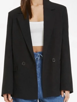 Svart oversized kavaj/kappa med knappar - Prislapp kvar! Stilren svart oversized kavaj med två knappar framtill och klassiska slag. Kavajen har raka axlar och en avslappnad passform, perfekt att slänga över en t-shirt eller topp. Tillverkad i ett mjukt tyg som ger en snygg och modern look.