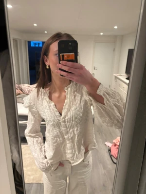 Vit romantisk blus med spetsdetaljer - Söt vit blus med långärmad design, volang vid axlarna och broderade spetsdetaljer. Blusen har knappar framtill. Säljer pga dålig användning!