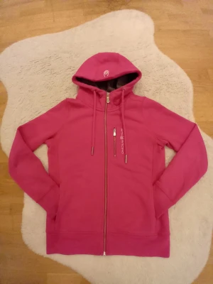 Rosa hoodie från Sail racing - Snygg rosa hoodie från sail racing med dragkedja framtill och en praktisk bröstficka med zip. Hoodien har justerbar huva med snören och mjukt grått foder på insidan av huvan. Perfekt för en avslappnad och sportig stil.