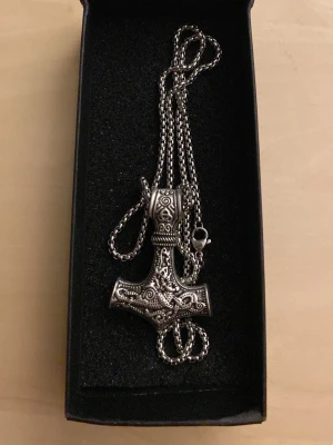 Vikinginspirerat Thorshammare i stål halsband 55cm - Säljer ett vikinginspirerat halsband med en detaljerad Thorshammare i antiksilverfärgad metall. Hänget har snygga mönster och kedjan är grov och matchar stilen perfekt. Perfekt accessoar för dig som gillar nordisk mytologi och vill sticka ut.
