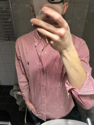 Rödvit randig skjorta från Ralph Lauren - Klassisk röd och vit randig skjorta från Ralph Lauren med button-down krage och broderad logga på bröstet. Skjortan har lång ärm, knappar framtill och är tillverkad i bomull. Perfekt för en clean och stilren look. Storlek M men sitter tajt så perfekt för S också.