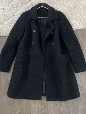 Svart rock med dubbelknäppning - Stilren svart rock med dubbelknäppning och bred krage. Två snedställda fickor framtill och helsvart foder på insidan. Klassisk rak passform som funkar till många olika looks. Storlek som en S