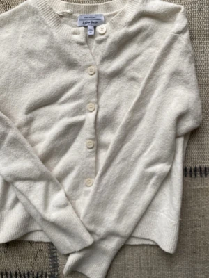 Vit kofta från & Other Stories - Klassisk vit kofta från & Other Stories Paris Atelier med rund halsringning och fyra knappar framtill. Tillverkad i mjukt material med ribbade muddar vid ärmslut och nederkant. Perfekt att slänga över en topp för en enkel och clean look.