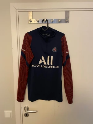PSG Tröja  - Snygg långärmad träningströja från PSG i marinblått med röda ärmar och mönster. Tröjan har halv dragkedja, klubbmärke på bröstet och Nike-logga på ärmen. Tillverkad i ett lätt och funktionellt material, perfekt för fotbollsträning.
