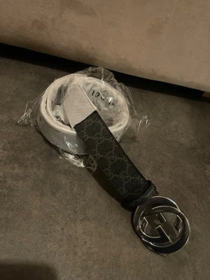 Svart Gucci bälte med GG-spänne - Snyggt svart bälte från Gucci med klassiskt GG-mönster längs hela remmen och ett stort, silverfärgat GG-spänne i metall. Perfekt accessoar för att lyfta vilken outfit som helst. Bältet är gjort i ett tåligt material med exklusiv känsla.