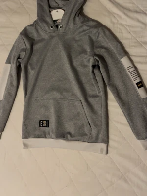 Grå hoodie med tryck på ärmen - Snygg grå hoodie från Softr med huva och vita muddar. Har en stor magficka och coolt tryck på ena ärmen samt en liten patch med texten 'EST' framtill. Perfekt för en avslappnad och trendig look. Tillverkad i mjukt material för extra komfort.