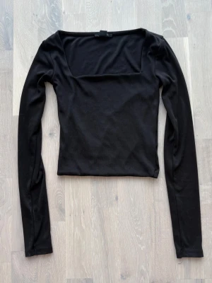 Svart långärmad topp med fyrkantig ringning - Stilren svart topp med lång ärm och fyrkantig ringning. Modellen är croppad och har en tight passform som framhäver siluetten. Perfekt att matcha med höga jeans eller kjol. Tillverkad i mjukt material för en bekväm känsla.