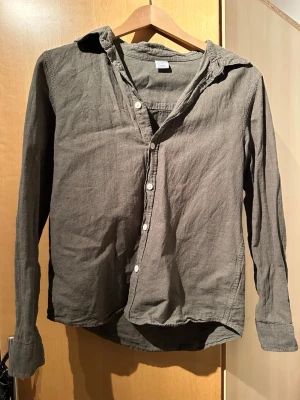 Grå linneskjorta med knappar - Snygg grå skjorta i linne med klassisk krage och vita knappar framtill. Skjortan har lång ärm och en avslappnad passform, perfekt för en stilren look. Enkel design utan mönster eller extra detaljer.