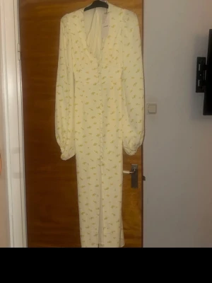 Ljusgul långklänning med blommor - Supersöt långklänning i ljusgul nyans med små blommiga detaljer. Klänningen har v-ringning, markerad midja och långa puffärmar. Knappar framtill hela vägen ner ger en vintagevibe. Perfekt för sommarens alla tillfällen!