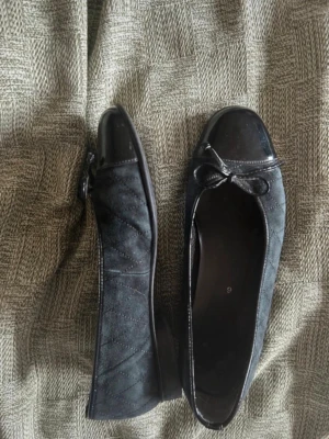 Svarta ballerinaskor med rosett - Snygga svarta ballerinaskor med lackad tå och liten rosett framtill. Skorna har en matt finish i mockaimitation och detaljer i blankt material. Klassisk rundad form och låg sula, perfekta för en stilren look.