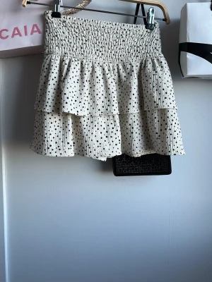 Prickig volangkjol i beige och svart - Supersöt kortkjol med volanger i beige med svarta prickar. Kjolen har smock i midjan för en bekväm passform och dubbla volanglager som ger en lekfull look. Perfekt för dig som vill ha en trendig och luftig kjol till sommaren.