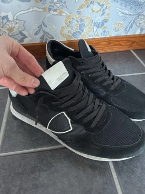 Svarta sneakers från Philippe Model - Snygga svarta sneakers från Philippe Model med vita detaljer och svart sula. Skorna har en mix av mocka och textil på ovandelen samt vita inslag på hälen och tungan. Klassisk låg modell med svarta skosnören och bekväm innersula.