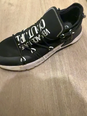 Svarta sneakers Versace Jeans Couture - Snygga svarta sneakers från Versace Jeans Couture med vita detaljer och logga på både plös och sula. Skorna har en modern design med unika snörningar och robust vit sula. Perfekta för dig som vill sticka ut med ett exklusivt streetwear-utseende.