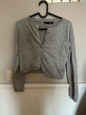 Grå cropped kofta från 157 - Snygg grå cropped kofta från 157 med rund hals och knappar framtill. Koftan har långa ärmar och ribbade muddar vid hals, ärmslut och nederkant. Perfekt att slänga över en t-shirt eller linne för en chill look.