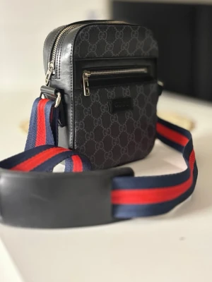 Gucci svart axelväska med GG-mönster - Snygg svart axelväska från Gucci med klassiskt GG-mönster i grått. Väskan har en framficka med silverfärgad dragkedja och bred axelrem i blått och rött. Tillverkad i slitstarkt canvasmaterial med läderdetaljer. Perfekt för dig som vill ha en trendig och praktisk väska.