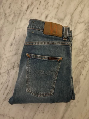 Nudie jeans - Säljer ett par klassiska blå raka jeans från Nudie Jeans med snyggt slitna detaljer och kontrastsömmar. Jeansen har fem fickor, normal midja och är tillverkade i bomull. Perfekta för en avslappnad och trendig look.