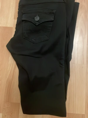 Svarta Joey flare jeans True Religion - Säljer ett par svarta Joey Low Rise Flare jeans från True Religion. De har låg midja, utsvängda ben och en snygg ficka med lock och knapp bak. Perfekta för dig som gillar en cool och trendig look. Oxå helt nya hann inte lämna tillbaka!