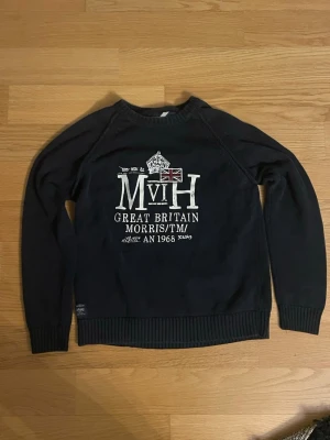 Mörkblå tröja från Morris - Mörkblå stickad tröja från Morris med vit tryckt text och brittisk flagga på bröstet. Tröjan har rund hals, ribbade muddar och en patch med Morris-logga vid ärmslutet. Perfekt för dig som gillar klassisk brittisk stil.