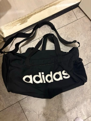 Svart sportbag från Adidas - Säljer en svart sportbag från Adidas med stort vitt logotryck på sidan. Väskan har både bärhandtag och justerbar axelrem samt meshficka på sidan. Perfekt för gymmet eller träningen, rymmer mycket och är lätt att bära.