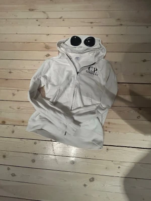 Vit hoodie från C.P. Company med goggles - Säljer en vit hoodie från C.P. Company med dragkedja och ikoniska goggles i huvan. Hoodien har fickor framtill och logga på bröstet. Perfekt för dig som vill sticka ut med en streetwear-look.Den är använd ett fåtal gånger.nypris runt 2000