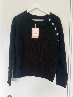 Svart stickad tröja med blommiga knappar - Snygg svart stickad tröja från Design Only med dekorativa blommiga knappar på axeln. Tröjan har rund halsringning, långa ärmar och ribbade muddar. Perfekt för dig som gillar en enkel men ändå unik look.