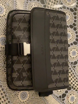 Svart axelväska från Lacoste - Snygg svart axelväska från Lacoste med geometriskt mönster i grått. Väskan har justerbar axelrem, metallspänne och präglad Lacoste-logga på framsidan. Tillverkad i slitstarkt syntetmaterial och har en modern, rektangulär form.