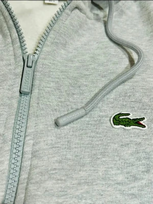 Grå zip hoodie från Lacoste - Snygg ljusgrå hoodie från Lacoste med dragkedja, huva och klassiska krokodilloggan broderad på bröstet. Tillverkad i mjuk bomullsblandning med dragsko i huvan och ribbade muddar. Perfekt för en chill och stilren look.