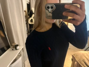 Mörkblå kabelstickad tröja Polo Ralph Lauren - Mörkblå kabelstickad tröja från Polo Ralph Lauren med den klassiska röda loggan broderad på bröstet. Tröjan har rund halsringning och långa ärmar. Perfekt för dig som gillar en tidlös och stilren look.