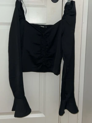 Svart croppad topp med snörning - Trendig svart croppad topp med långa ärmar och snörning framtill. Toppen har vida ärmslut och en lätt v-ringad urringning. Perfekt för dig som vill ha en edgy och stilren look.