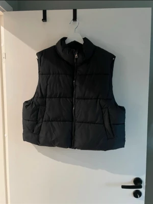 Svart dunväst från 157, storlek XL - Stilren svart dunväst från 157 med hög krage och dragkedja framtill. Västen har två stora fickor på sidorna och quiltad design som ger en schysst look. Perfekt att slänga över en hoodie eller tröja när det är kyligt ute.