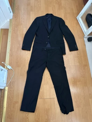 Svart kostym  från glory days - Stilren svart kostym med klassisk kavaj och matchande byxor. Kavajen har två knappar framtill, fickor med lock och smal krage. Byxorna är raka.” Ny pris 1600” mitt pris 420