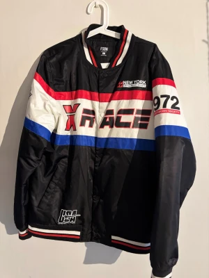 Svart bomberjacka X RACE - Snygg svart bomberjacka från FSBN med racing-inspirerad design. Jackan har röda, vita och blå detaljer, tryck med 'X RACE' och 'NEW YORK RACING SOCIETY' samt randiga muddar. Materialet är glansigt och jackan har tryckknappar framtill.