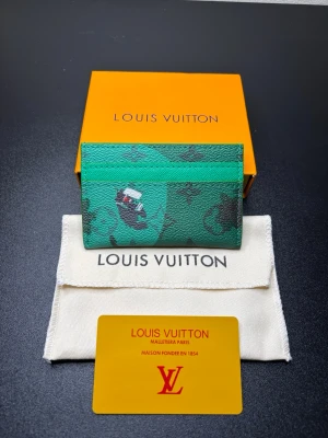 Grön Louis Vuitton korthållare - Snygg grön korthållare från Louis Vuitton med klassiskt monogrammönster och en unik illustration på framsidan. Tillverkad i tåligt canvasmaterial och har plats för flera kort. Perfekt accessoar för dig som vill ha något stilrent och trendigt.