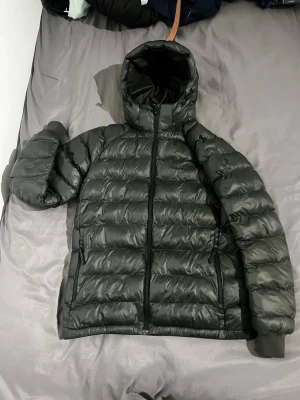 Svart dunjacka med huva - Snygg svart dunjacka med huva och dragkedja framtill. Jackan har två sidofickor med dragkedja och ribbade muddar vid ärmsluten. Perfekt för kalla vinterdagar och har en klassisk, puffig design.