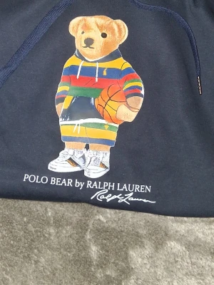 Mörkblå Polo Bear hoodie Ralph Lauren - Mörkblå hoodie från Polo Ralph Lauren med ikoniska Polo Bear-trycket på bröstet. Björnen har färgglad randig hoodie och sneakers, samt håller en basketboll. Klassisk huva med dragsko och mjukt material. Perfekt för en avslappnad streetstyle-look.
