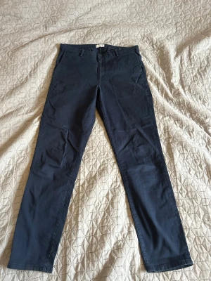 Mörkblå chinos med smal passform - Snygga mörkblå chinos med klassisk design. Byxorna har framfickor, bälteshällor och två bakfickor med en knapp. Perfekta för en stilren och avslappnad look. Materialet är mjukt och skönt.