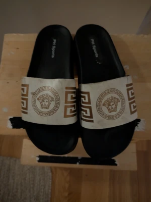 Svarta sandaler från PH Sports - Svarta slip-in sandaler från PH Sports med bred vit rem över foten. Remmen har guldfärgade grekiska mönster och ett lejonhuvud i mitten. Snygg kontrast mellan svart och vitt, och coolt mönster som sticker ut. Perfekta för chill dagar eller till stranden.