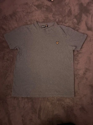 Grå t-shirt från Lyle & Scott - Snygg grå t-shirt från Lyle & Scott med klassisk gul broderad örn på bröstet. T-shirten har rund hals och korta ärmar, perfekt basic-plagg till garderoben. Tillverkad i mjuk bomull för skön känsla.