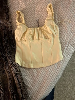 Gul linnetopp med rynkade axelband - Söt ljusgul linnetopp med breda, rynkade axelband och lätt croppad passform. Toppen har en mjuk, något glansig yta och paneler framtill som ger en strukturerad look. Perfekt för varma dagar och enkel att styla till jeans eller kjol.