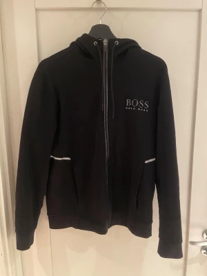 Svart zip hoodie från Hugo Boss - Snygg svart hoodie med dragkedja från Hugo Boss. Har huva med drag, vita detaljer vid fickorna och logga på bröstet. Perfekt för en sportig eller stilren outfit!  Modellen är 185 cm. 