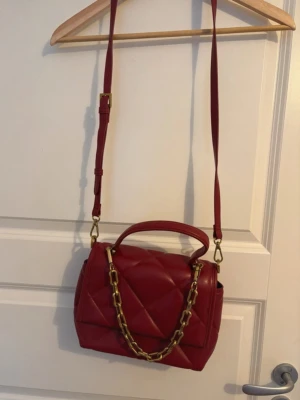 Röd quiltad handväska med kedja - Snygg röd handväska i quiltat mönster med justerbar axelrem och guldfärgad kedjedetalj. Väskan har ett topphandtag och är tillverkad i konstläder. Perfekt accessoar för att lyfta din outfit. Kolla min profil för fler plagg!