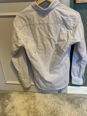 Randig skjorta med knappar - Klassisk skjorta med smala blå och vita ränder, knappar framtill och button-down krage. Långärmad modell i mjuk bomull som passar perfekt till jeans eller chinos. Enkel och stilren design som funkar till många olika looks.