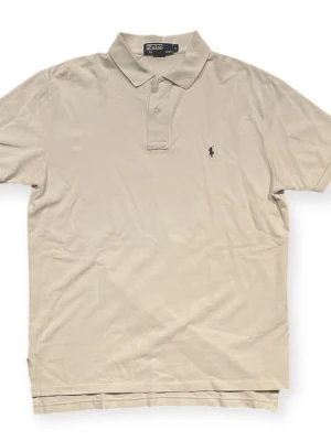 Beige pikétröja från Polo Ralph Lauren - Klassisk beige pikétröja från Polo Ralph Lauren med korta ärmar och liten broderad logga på bröstet. Tröjan har krage och knappar vid halsen. Perfekt till en avslappnad men ändå stilren look.