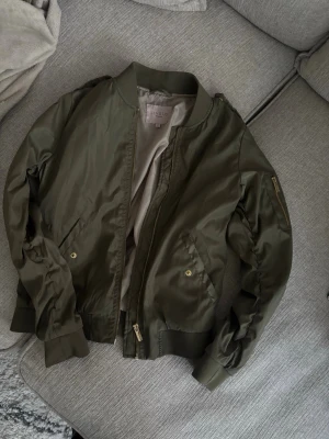 Olivgrön bomberjacka från Livelo - Snygg olivgrön bomberjacka från Livelo med guldfärgade dragkedjor och fickor. Jackan har klassisk ribbad krage, muddar vid ärmslut och nederkant samt en ärmficka med dragkedja. Materialet är glansigt och ger en modern look.