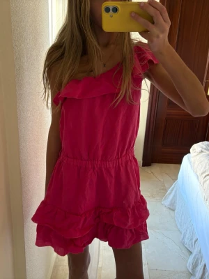 Rosa one shoulder volangklänning - Säljer en superfin rosa kortklänning med oneshoulder och volangdetaljer. Klänningen har dubbla volanger nedtill för en extra söt look. Perfekt för dig som vill sticka ut med en färgstark och trendig stil. 💓