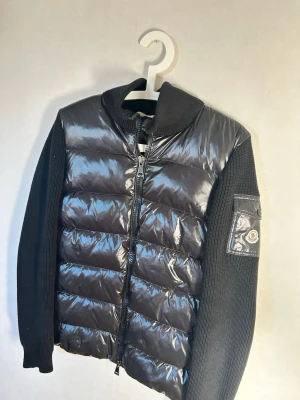 Svart cardiganjacka från Moncler - Snygg svart cardiganjacka från Moncler med glansig vadderad front och stickade ärmar. Jackan har dragkedja framtill, hög krage och Moncler-logga på ärmen. Perfekt för lager-på-lager och streetstyle.